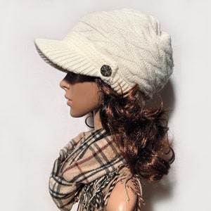 May include: A white knitted beanie with a button detail and a brim. The beanie is worn on a mannequin with brown hair and a plaid scarf.