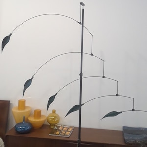 May include: A black metal mobile sculpture with five green leaf-shaped pieces hanging from curved arms. The mobile is suspended from a black metal rod and is displayed on a wooden table with three vases.