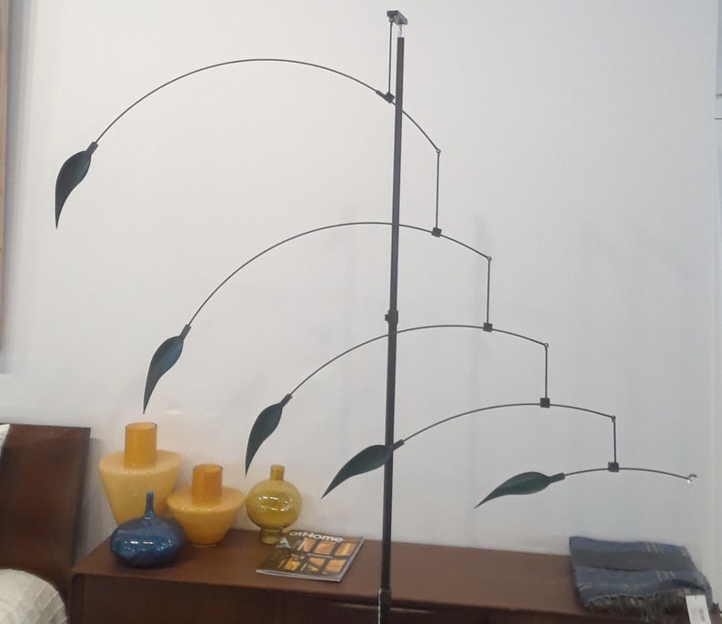 May include: A black metal mobile sculpture with five green leaf-shaped pieces hanging from curved arms. The mobile is suspended from a black metal rod and is displayed on a wooden table with three vases.