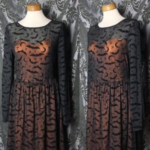 May include: Black long-sleeved dress with a bat, crescent moon, and cross pattern. The dress has a round neckline and a fitted bodice with a flared skirt. The dress is displayed on a mannequin.