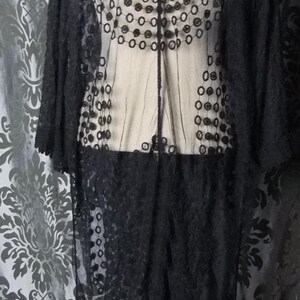 May include: A long, black, sheer lace garment with a floral and circular pattern. The garment has long sleeves and a decorative back panel. The lace is detailed and the garment is displayed on a mannequin.
