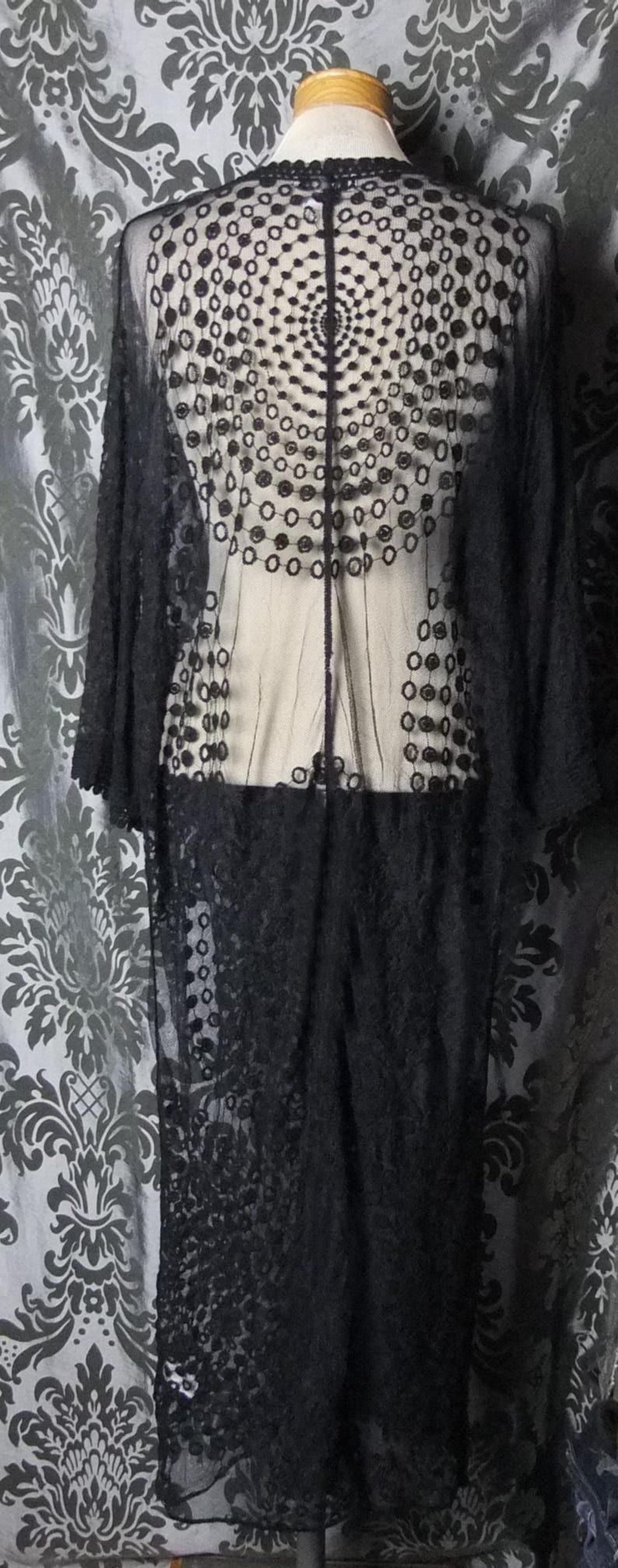 May include: A long, black, sheer lace garment with a floral and circular pattern. The garment has long sleeves and a decorative back panel. The lace is detailed and the garment is displayed on a mannequin.
