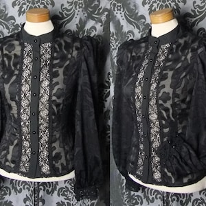 May include: Black sheer blouse with a high collar and long, puffed sleeves. The front features a vertical panel of white lace and black buttons. The blouse is displayed on a mannequin, showcasing its intricate floral pattern and vintage style.