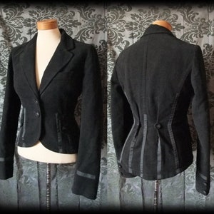 May include: A black fitted blazer featuring a notched lapel, long sleeves with cuff buttons, and a front button closure. The back displays a structured peplum-style design with vertical ribbon detailing and a central button accent at the waist.