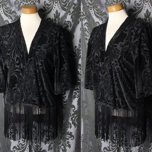 May include: Black velvet kimono with a V-neck and short sleeves. The kimono features a tonal, ornate pattern and a fringe hemline. The garment is displayed on a mannequin.
