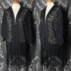 May include: A black kimono jacket with a floral pattern and fringe trim. The sheer fabric is draped over a mannequin. The jacket has short sleeves and a V-neck. The fringe hangs from the bottom hem, adding a decorative element.