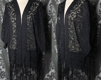 May include: A black, open-front kimono-style jacket with a floral pattern and fringe trim. The sheer fabric drapes over a mannequin, showcasing the design. The jacket's sleeves are short, and the fringe hangs from the bottom hem.