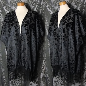 May include: Black velvet kimono with a floral pattern and fringe trim. The open-front design drapes elegantly, showcasing the intricate detailing of the fabric. This garment is a stylish addition to any wardrobe.