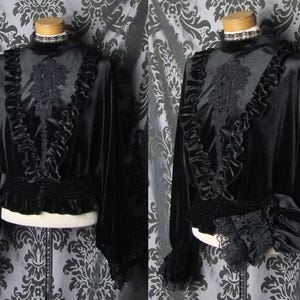 May include: A black velvet blouse with long sleeves, a high collar, and ruffled trim. The blouse features a decorative lace panel on the chest and lace cuffs. The waist is cinched with a gathered elastic band. The garment is displayed on a mannequin.