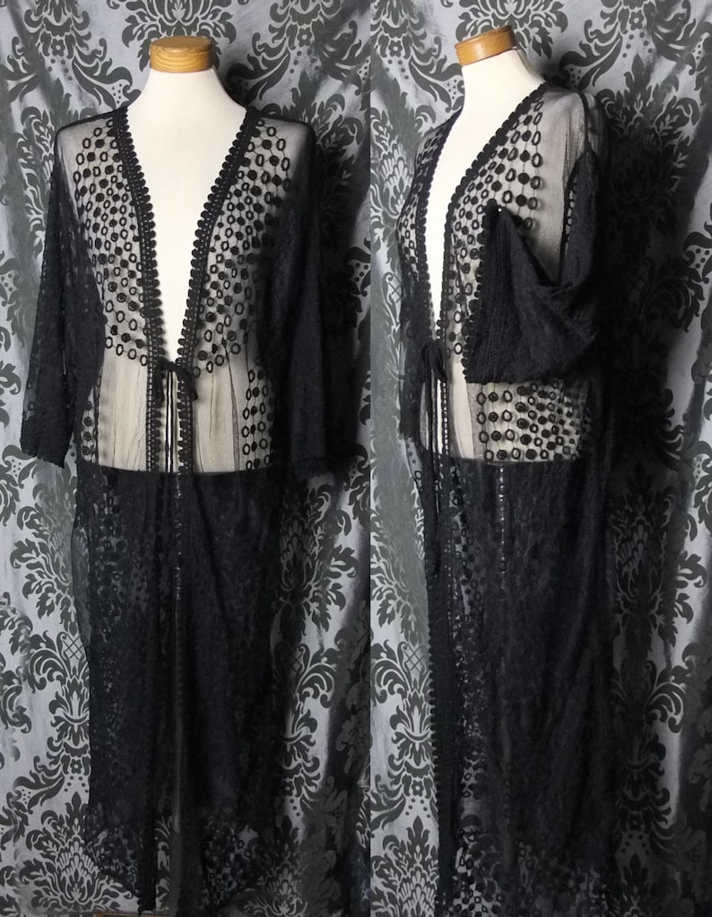 May include: Black lace duster coat with a V-neck and long sleeves. The sheer fabric features a dotted pattern and is trimmed with a scalloped edge. The coat is displayed on a mannequin.
