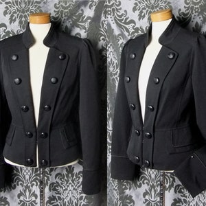 Puede incluir: Chaqueta negra con cuello alto y cierre de botones cruzados. La chaqueta presenta una cintura entallada, pequeños bolsillos y mangas largas con puños abotonados. La prenda se exhibe en un maniquí.