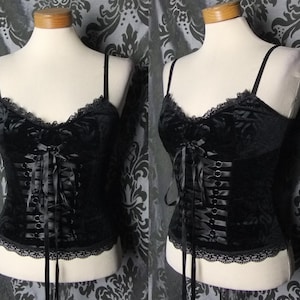 May include: A black velvet corset top featuring a floral pattern, thin shoulder straps, and a front lace-up closure with black ribbon. The garment is finished with delicate black lace trim along the neckline and the bottom hem.