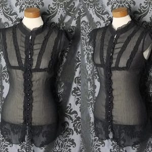 May include: A black sheer sleeveless blouse featuring a high ruffled collar, a vertical row of small round buttons down the center front, and decorative ruffled trim accents along the bodice and armholes, displayed on a mannequin torso.