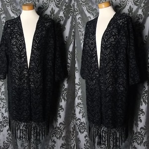 May include: Black open-front kimono-style jacket with a patterned design and fringe trim. The jacket is displayed on a mannequin. The fabric appears to be a sheer material, and the design is visible throughout the garment.