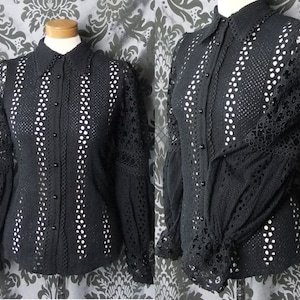 May include: Black button-down blouse with a collared neckline and long sleeves. The blouse features intricate cut-out detailing and a scalloped trim on the sleeves and cuffs. The sleeves are puffy at the shoulders and fitted at the wrists.