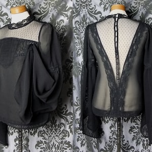 May include: Black blouse with sheer sleeves and a lace-trimmed neckline. The front features a decorative panel with pleats and beaded accents. The back has a sheer panel with a lace trim and button closure. The blouse is displayed on a mannequin.