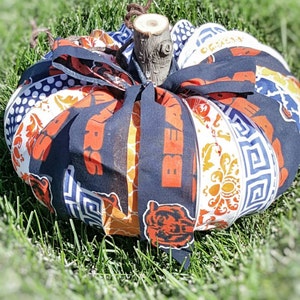 May include: A fabric pumpkin decorated with a patchwork of different patterns, including a blue and white geometric pattern, a white and orange floral pattern, and a navy blue fabric with the words "Bears" in orange letters. The pumpkin is tied with a navy blue ribbon and has a brown wooden stem.