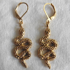 Könnte beinhalten: Ein Paar goldene Schlangenohrringe mit strukturierter Oberfläche. Die Ohrringe haben einen Bügelverschluss.