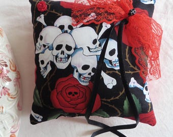 May include: A small, square pillow with a black background featuring white skulls, red roses, and crossbones. It is adorned with a red lace trim and black ribbons. A white rose and a decorative vase are also in the image.
