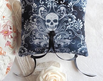 May include: A square, dark blue pillow with a white skull and ornate scrollwork design. The pillow has black ribbon ties and a small black rose. A white rose and a decorative ceramic vase are also in the image.