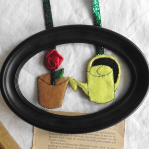 Può includere: Una cornice ovale nera con un fiore di feltro rosso in un vaso di feltro marrone e un annaffiatoio di feltro giallo con un manico nero.
