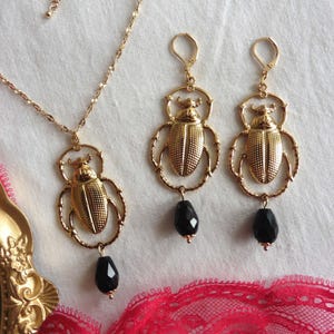 Op de afbeelding: Een goudkleurige sieradenset met een ketting en oorbellen. De ketting en oorbellen hebben een scarabee-ontwerp met een zwarte edelsteen hanger. De oorbellen hebben een oorhaaksluiting. De sieraden worden gepresenteerd op een witte ondergrond.