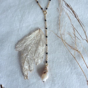 Peut inclure: Un collier en chaîne dorée avec des perles noires et un pendentif en forme de coquille. La coquille est blanche avec un accent doré.