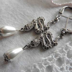 Pode incluir: Um par de brincos pendurados prateados. Cada brinco apresenta um design serpentino incrustado com pequenas pedras escuras e uma gota de pérola falsa branca em forma de pêra. Fechamento de alavanca.