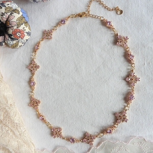 Może przedstawiać: Delikatny naszyjnik w kolorze złota z różowymi akcentami z kamieni szlachetnych. Naszyjnik zdobiony jest klastrami w kształcie kwiatów i mniejszymi, okrągłymi kamieniami szlachetnymi, tworzącymi błyszczący, kobiecy design. Naszyjnik jest prezentowany na białej powierzchni z koronkowym wykończeniem.