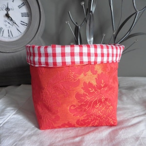 Peut inclure: Un panier de rangement doublé de tissu vichy rouge et blanc avec un motif floral. Le panier est fait d'un tissu doux et texturé.