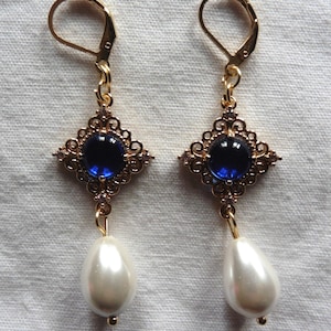 Peut inclure: Boucles d'oreilles pendantes dorées avec une pierre précieuse bleue et une perle blanche. Les boucles d'oreilles ont un design en filigrane.