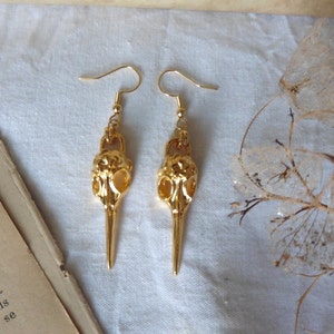 Puede incluir: Un par de pendientes de tono dorado con un diseño detallado de cráneo de pájaro. Los pendientes son colgantes y tienen un cierre de gancho.