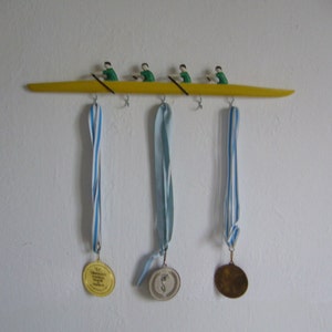 Puede incluir: Un colgador de medallas de pared de madera amarilla con forma de bote de remo con cuatro figuras con camisas verdes. Tres medallas con cintas azules y blancas cuelgan debajo. Una medalla es dorada, una plateada y otra de bronce.