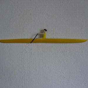 May include: A yellow wooden sculpture of a person rowing a boat. The boat is long and narrow, with the rower in a yellow outfit and a black oar. The sculpture is mounted on a white wall.