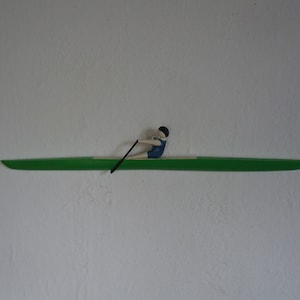 May include: A wooden wall decoration featuring a green kayak with a miniature figure of a rower in blue and black. The rower is positioned in the center of the kayak, holding a black oar. The background is a plain white wall.