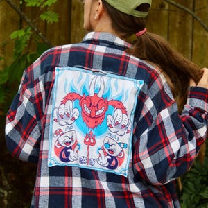 May include: A red, white, and blue plaid flannel shirt with a patch featuring cartoon characters. The patch depicts a red devil with blue flames and two cartoon cup characters. The shirt has a classic button-down design.