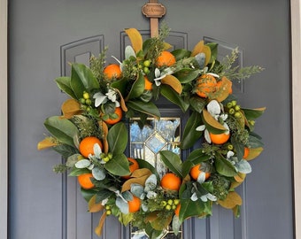 Könnte beinhalten: Ein Kranz aus Magnolienblättern und Grün mit orangefarbenen Fruchtakzenten. Der Kranz hängt an einer grauen Tür mit einem Messing-Willkommensschild.