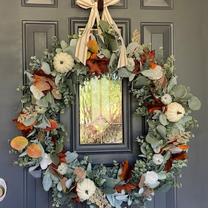 May include: A fall-themed wreath with white pumpkins, faux leaves in shades of orange, brown, and green, and eucalyptus greenery. The wreath is adorned with a striped ribbon and is hanging on a dark gray door.