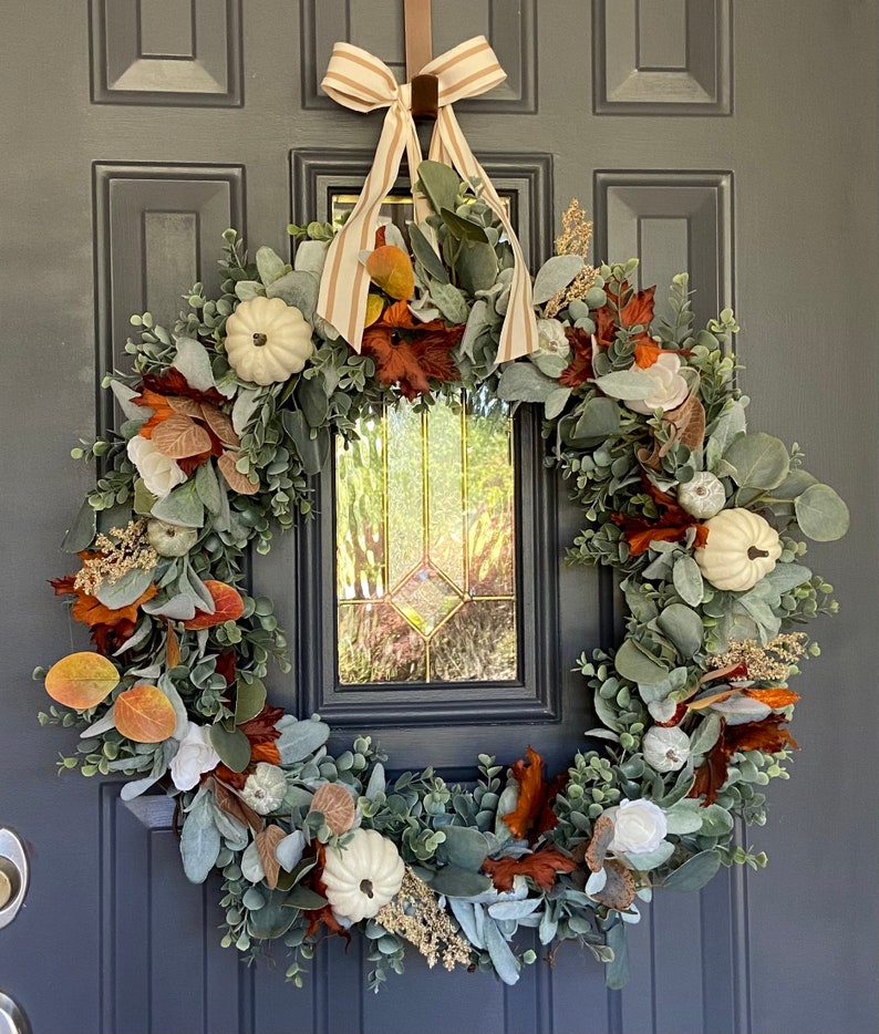 May include: A fall-themed wreath with white pumpkins, faux leaves in shades of orange, brown, and green, and eucalyptus greenery. The wreath is adorned with a striped ribbon and is hanging on a dark gray door.