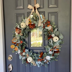 May include: A fall-themed wreath with white pumpkins, orange and brown leaves, and green foliage hangs on a dark blue door. A brown strap with the word "Welcome" is at the top, and a striped bow is tied at the top of the wreath.