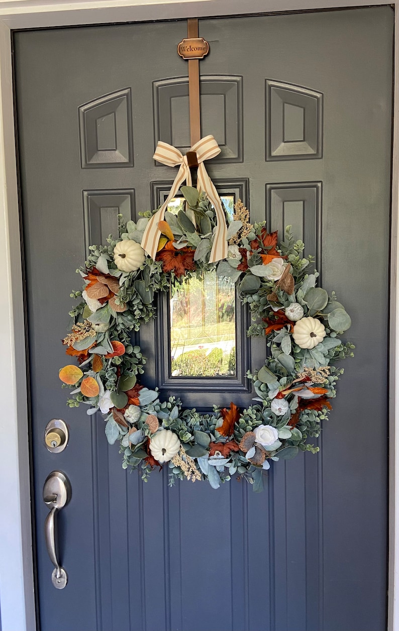 May include: A fall-themed wreath with white pumpkins, orange and brown leaves, and green foliage hangs on a dark blue door. A brown strap with the word "Welcome" is at the top, and a striped bow is tied at the top of the wreath.