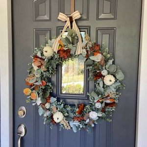 May include: A fall-themed wreath with white pumpkins, orange and brown leaves, and green foliage hangs on a dark gray door. A brown metal hanger with the word "Welcome" is above the wreath, and a striped bow is tied at the top.