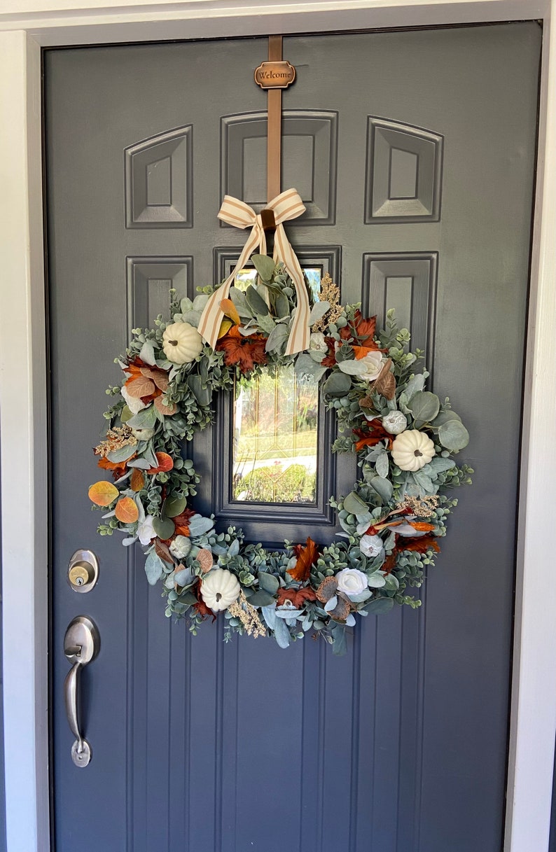 May include: A fall-themed wreath with white pumpkins, orange and brown leaves, and green foliage hangs on a dark gray door. A brown metal hanger with the word "Welcome" is above the wreath, and a striped bow is tied at the top.