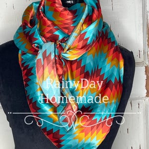 May include: A turquoise, red, and gold patterned silk bandana with a geometric design. The bandana is tied in a knot and is displayed on a mannequin. The text "Rainy Day Homemade" is visible on the bandana.