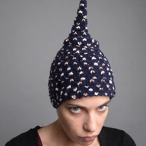 Può includere: Un berretto all'uncinetto blu navy scuro con una parte superiore appuntita a forma di cono. Il berretto presenta un motivo strutturato con piccole macchie bianche e color rame metallizzato sparse nel filato scuro, creando un aspetto maculato.