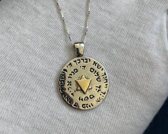 May include: A silver-toned pendant necklace featuring a circular pendant with Hebrew text and a gold-colored Star of David in the center. The pendant hangs from a delicate silver chain. The necklace is displayed against a light background.