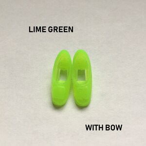 May include: A pair of lime green plastic doll shoes with a bow. The shoes have a cut-out design on the front.