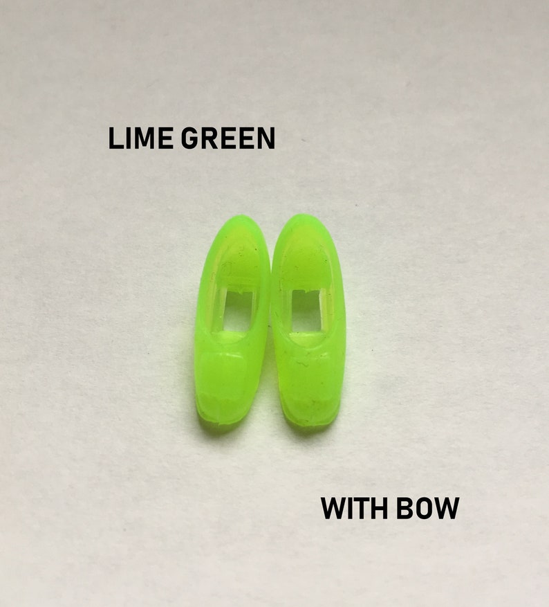 May include: A pair of lime green plastic doll shoes with a bow. The shoes have a cut-out design on the front.