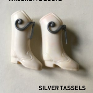 May include: A pair of white doll boots with silver tassels. The boots have a pointed toe and a low heel. The text "MAJORETTE BOOTS" and "SILVER TASSELS" is printed on the image.