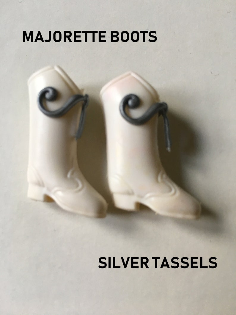 May include: A pair of white doll boots with silver tassels. The boots have a pointed toe and a low heel. The text "MAJORETTE BOOTS" and "SILVER TASSELS" is printed on the image.